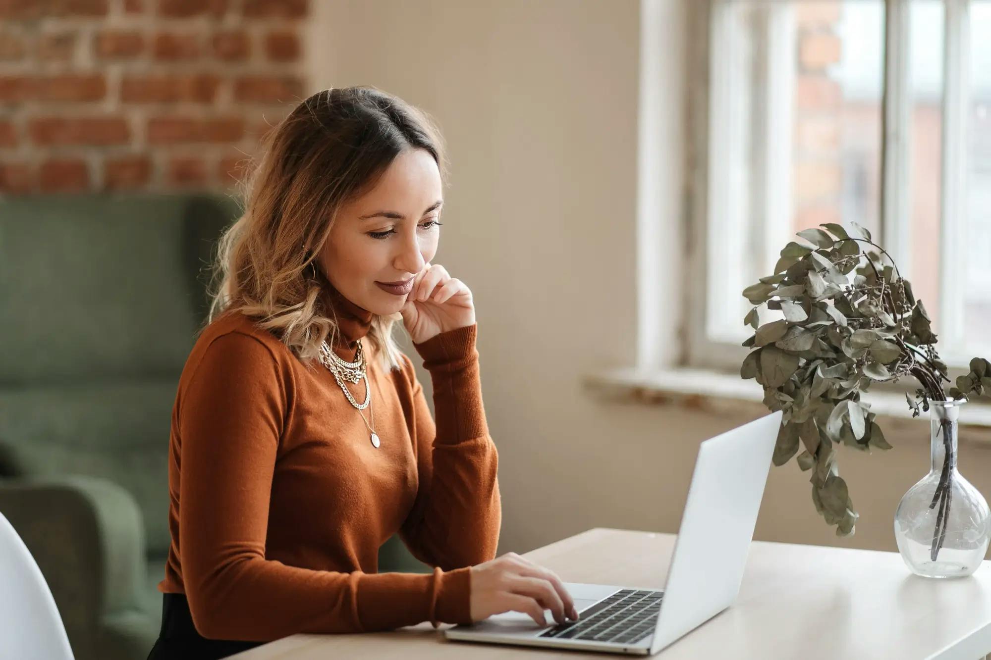 Woman in an orange sweater working on a laptop at a bright desk